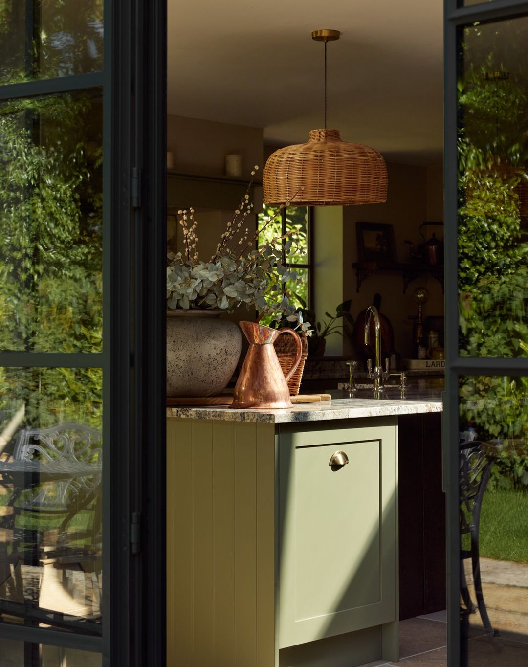 Interior view through open doors of a kitchen with a wicker pendant light, green cabinets, a stone countertop, and a copper pitcher.
