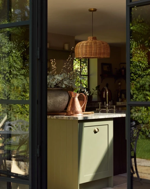 Interior view through open doors of a kitchen with a wicker pendant light, green cabinets, a stone countertop, and a copper pitcher.