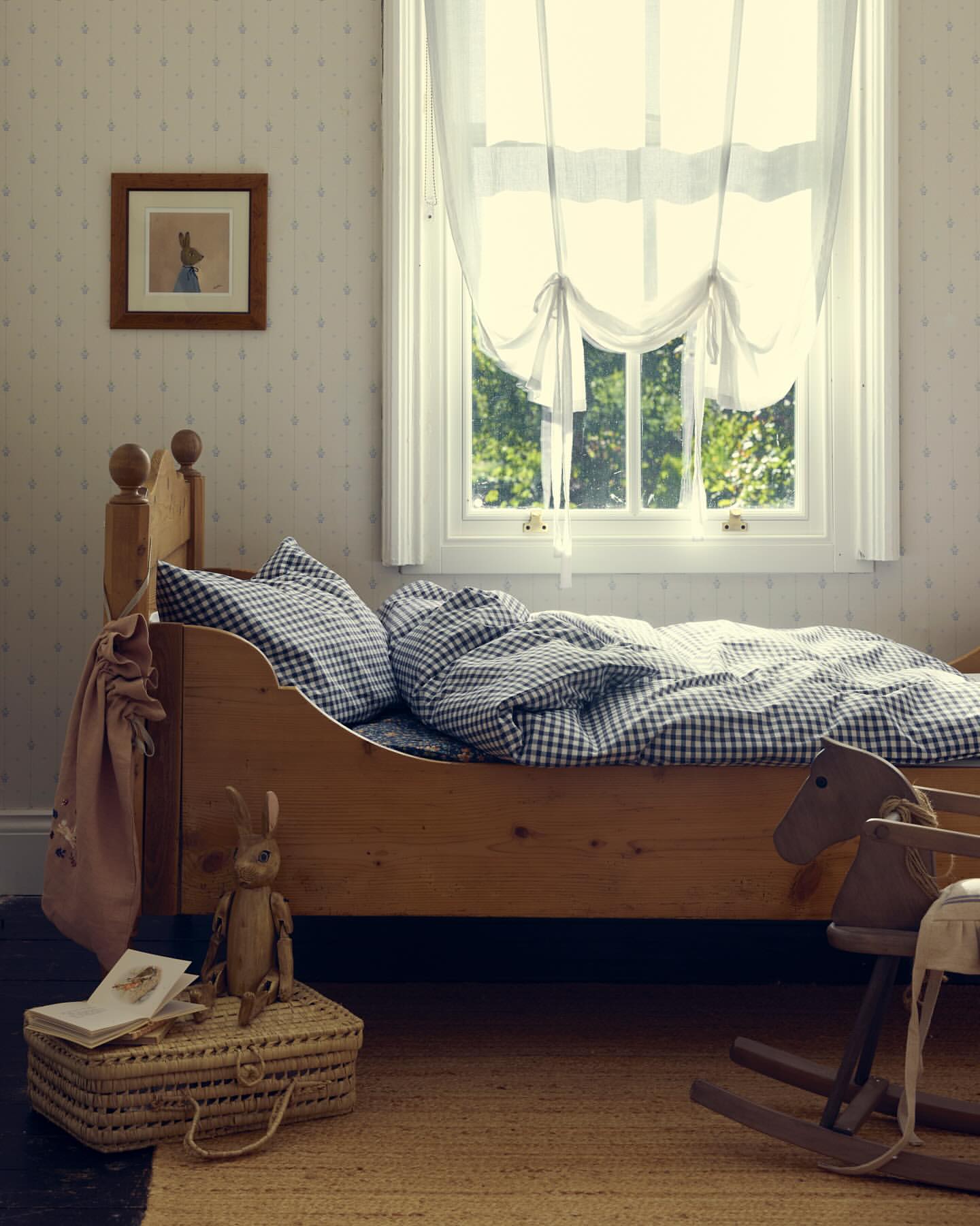 A child's bedroom with a wooden bed, a rocking horse, a toy rabbit, and a window with sheer curtains, all in soft, natural light.