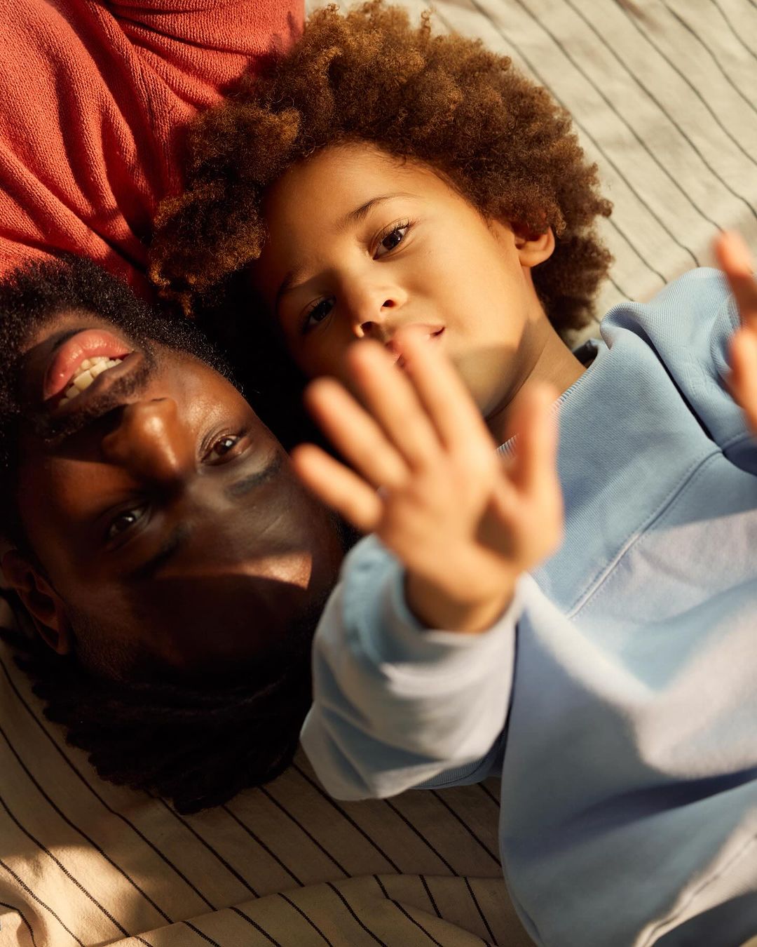 Father and child with curly hair lie on striped surface, smiling at the camera. Soft lighting, indoor setting, casual clothing.
