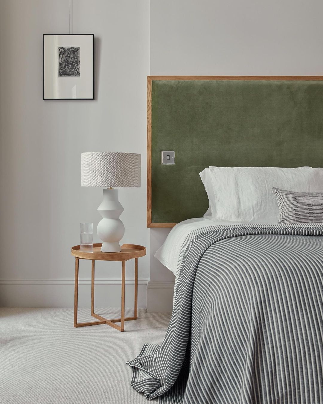 Bedroom interior featuring a bed with a striped blanket, green headboard, a bedside table with a lamp and glass, and framed artwork.