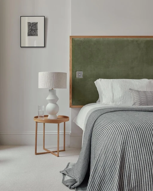 Bedroom interior featuring a bed with a striped blanket, green headboard, a bedside table with a lamp and glass, and framed artwork.