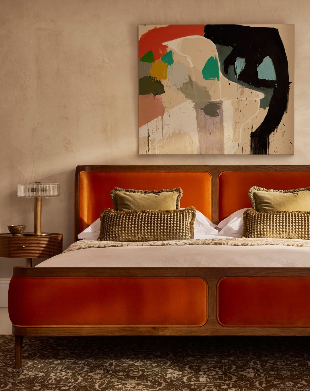 Interior bedroom shot: upholstered bed with pillows and side table with lamp under abstract art against neutral wall.