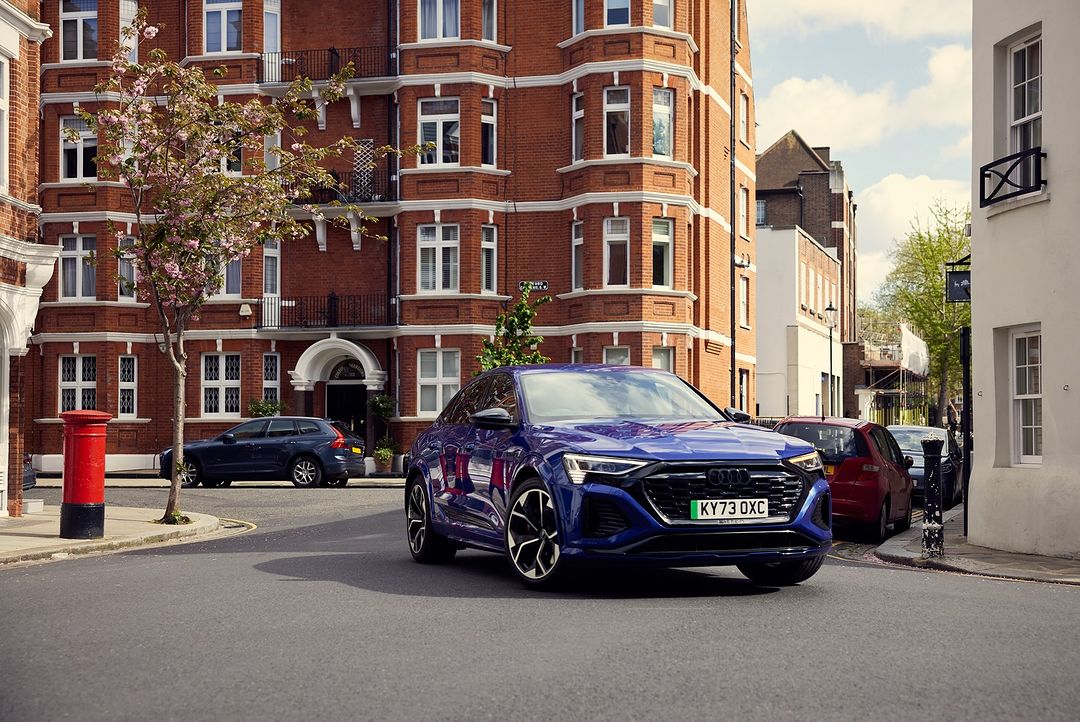 A blue Audi e-tron SUV on a city street, parked near red brick buildings and a traditional red British telephone box on a sunny day.