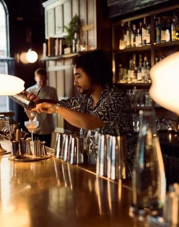 Bartender pouring a cocktail in a dimly lit bar with various bottles on shelves in the background, creating a vintage vibe.