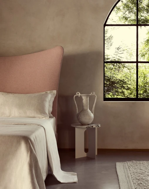 Bedroom interior showcasing a bed with linen bedding and a vase on a side table, viewed through a large window with a natural outdoor view.