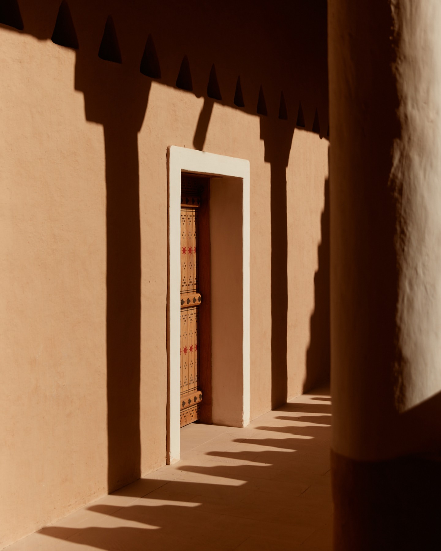 Architectural shot of a tan building exterior with a decorated wooden door, columns, geometric shadows, and a neutral, earthy color palette.