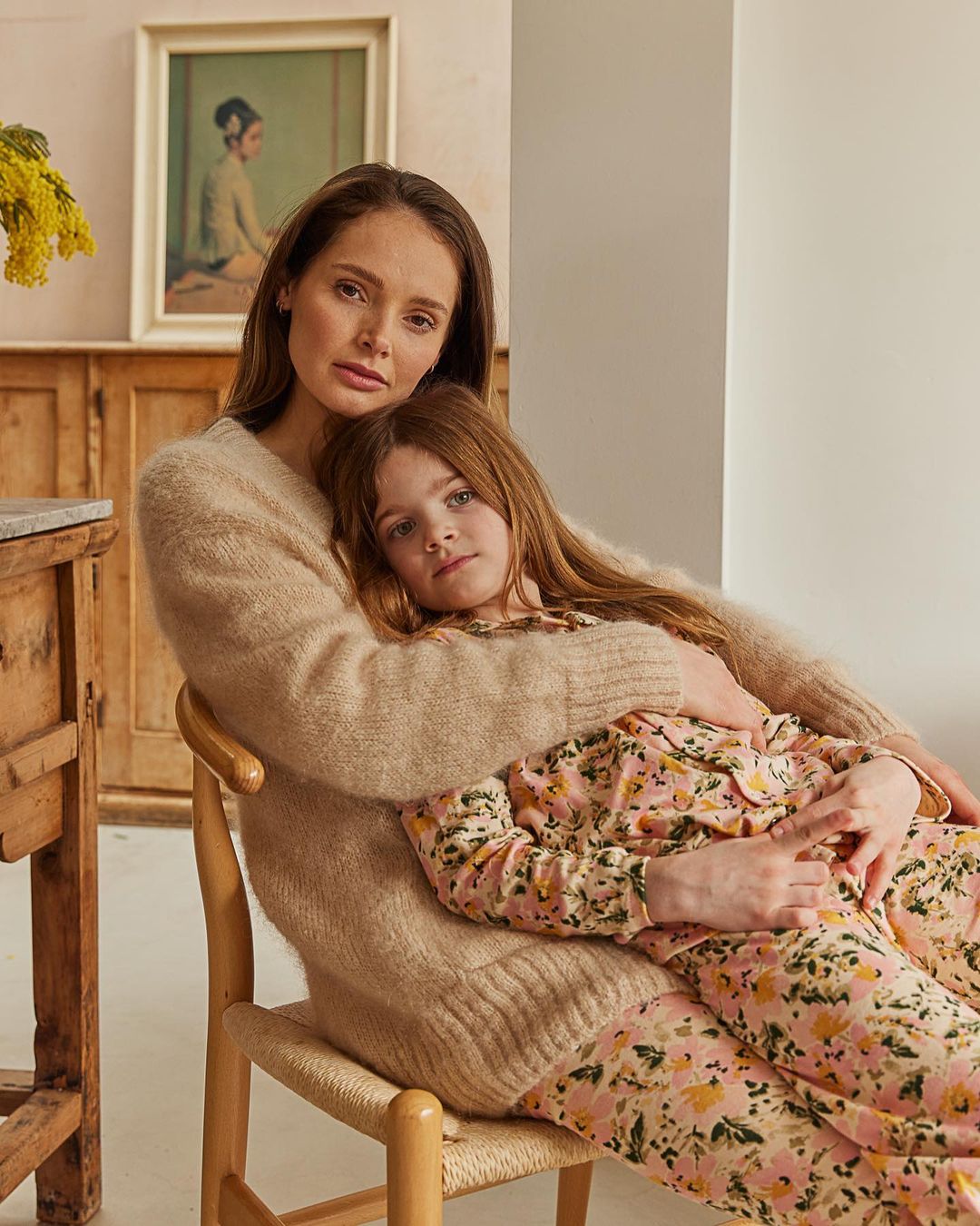 Woman and child pose indoors. Adult female sits on a chair with child in lap, embracing her. They wear soft sweaters and patterned pants.