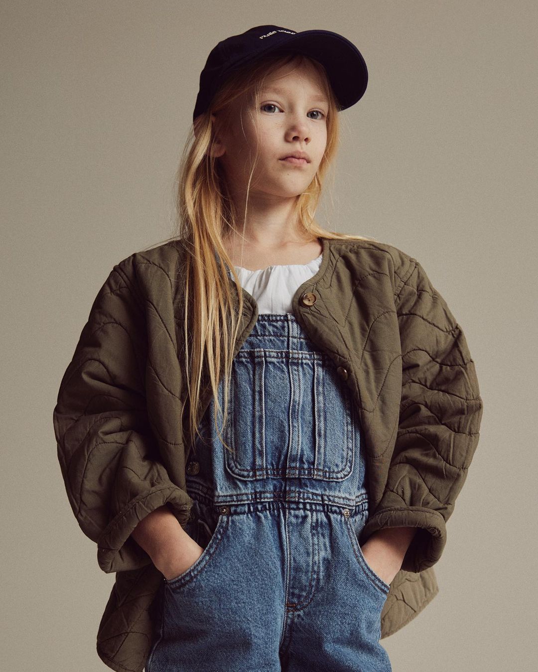Portrait of a young girl wearing a navy baseball cap, olive-green quilted jacket, and denim overalls against a neutral backdrop.