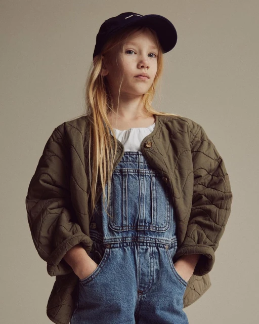 Portrait of a young girl wearing a navy baseball cap, olive-green quilted jacket, and denim overalls against a neutral backdrop.