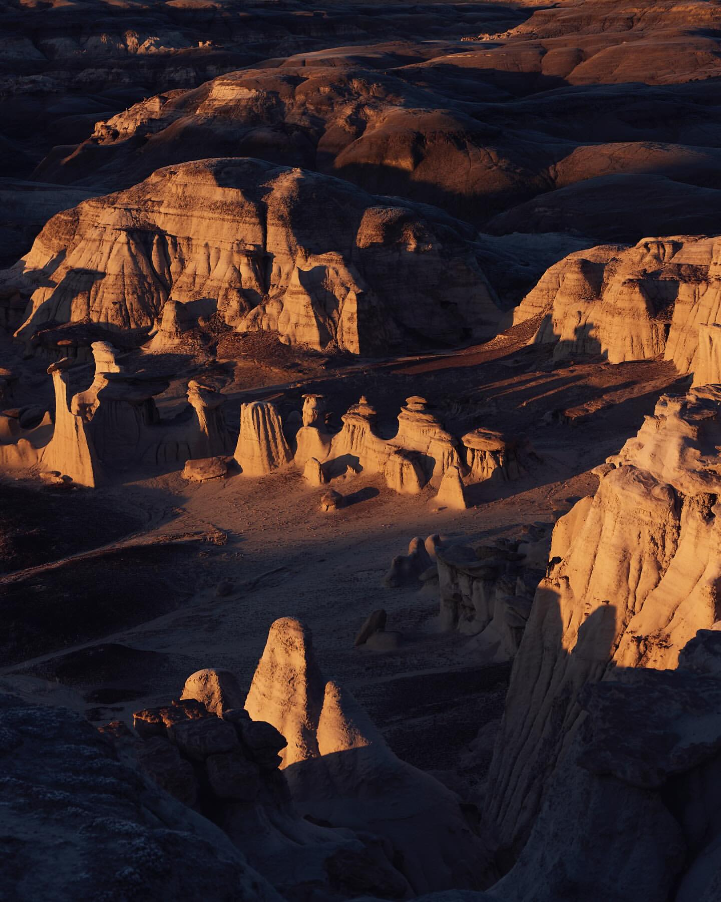 Dramatic golden light illuminates rock formations in a desert landscape. Eroded hoodoos and mesas create a stark, beautiful scene.