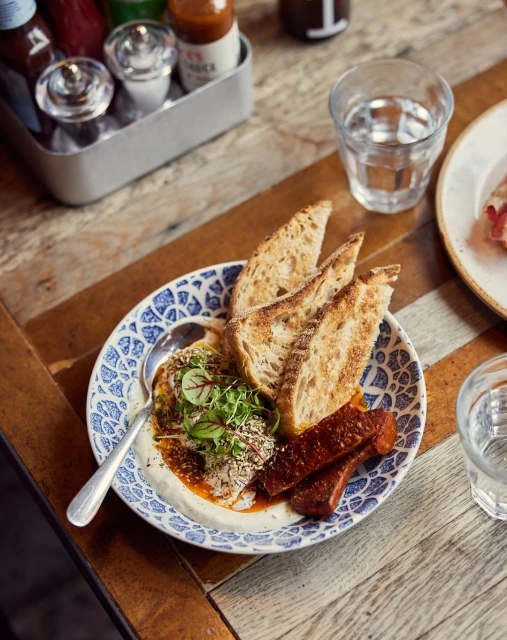 Overhead shot of a blue patterned plate with a breakfast meal of bread, sausage, sauce, and herbs on a wooden table with drinks.
