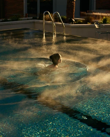 A child swims in an outdoor swimming pool with steam rising from the water, captured in golden light during late afternoon.