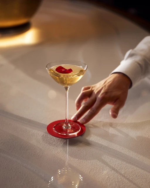 Cocktail glass with light-colored liquid, a red flower petal, red coaster, hand, and white shirt sleeve on a gray surface.