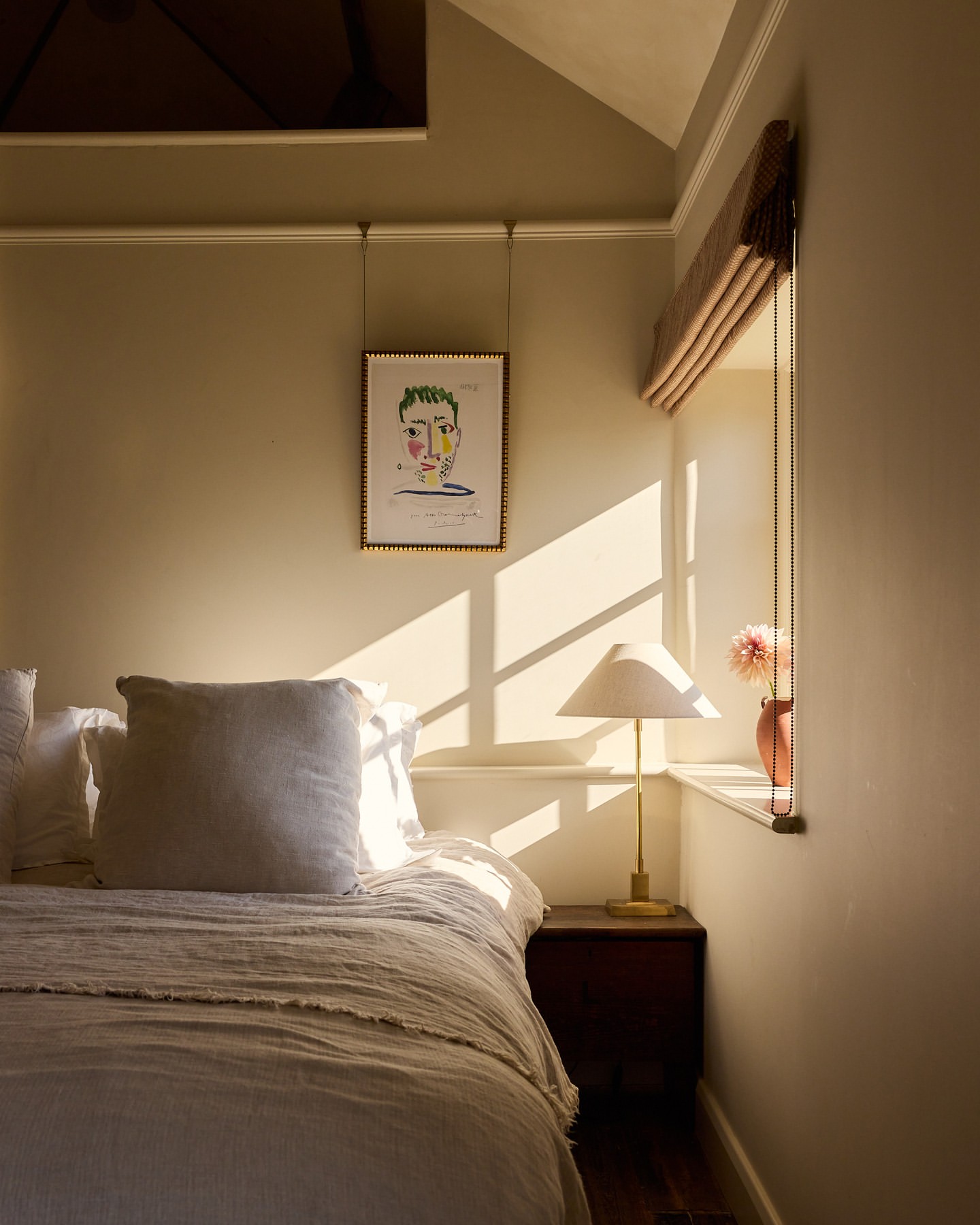 Bedroom interior with a bed, nightstand, lamp, window with sunlight, and wall art in a neutral color palette creating a peaceful setting.
