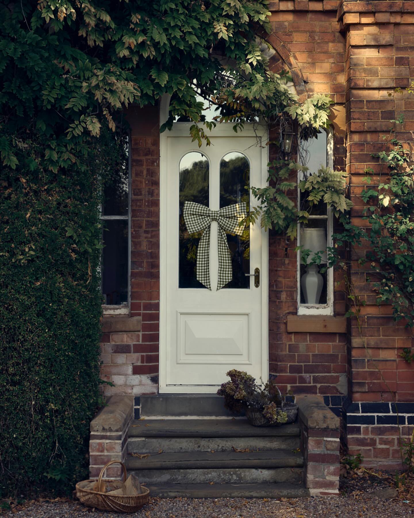 Exterior of a brick house featuring a white door adorned with a bow. Lush greenery surrounds the doorway, enhancing the home's charm.
