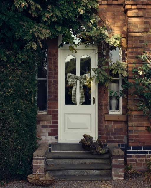 Exterior of a brick house featuring a white door adorned with a bow. Lush greenery surrounds the doorway, enhancing the home's charm.