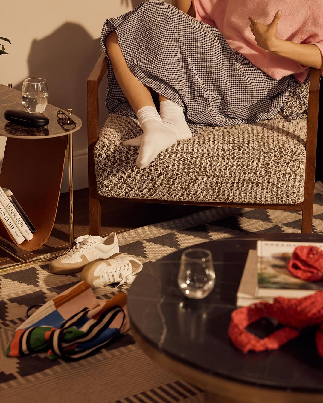 Woman in a chair with legs visible, surrounded by shoes, books, and glasses on nearby tables and patterned rug in a warm interior.