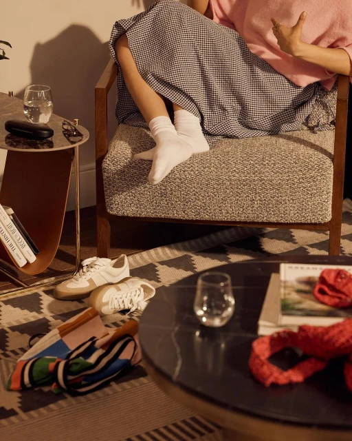Woman in a chair with legs visible, surrounded by shoes, books, and glasses on nearby tables and patterned rug in a warm interior.