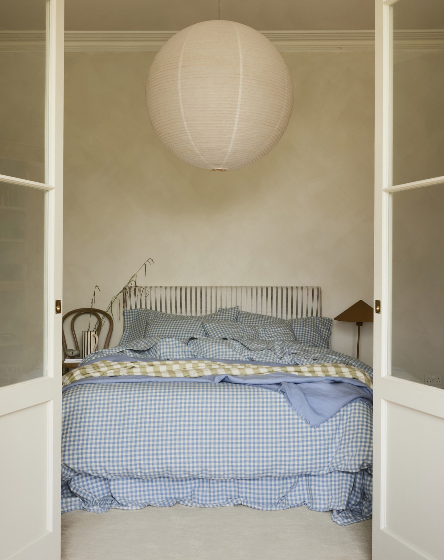 Bedroom interior with a gingham-patterned bed, a white paper lantern hanging from the ceiling, and white double doors framing the scene.