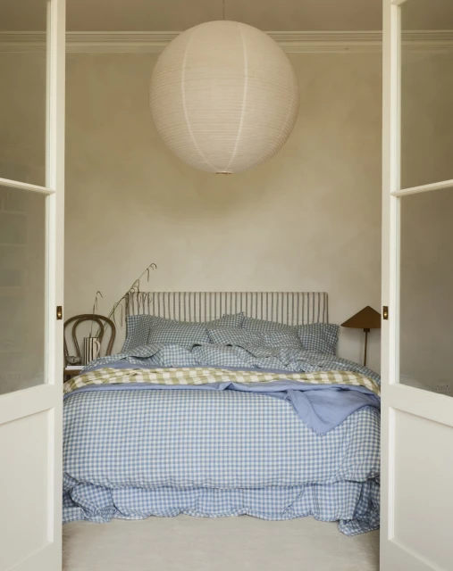 Bedroom interior with a gingham-patterned bed, a white paper lantern hanging from the ceiling, and white double doors framing the scene.