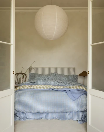 Bedroom interior with a gingham-patterned bed, a white paper lantern hanging from the ceiling, and white double doors framing the scene.