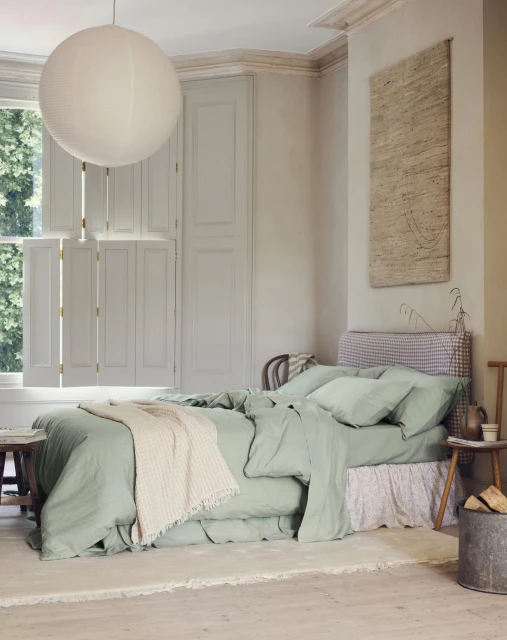 Bedroom interior with a bed dressed in green linen, a white blanket, and a round paper lantern hanging from the ceiling.