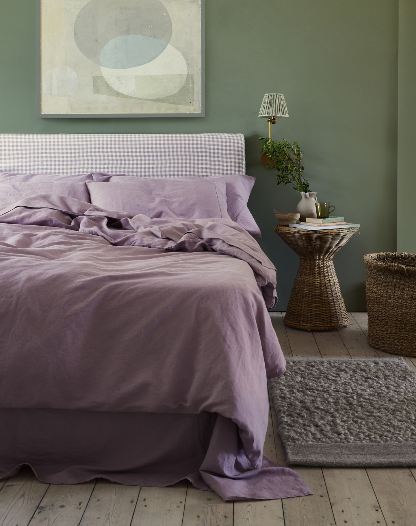 Bedroom interior with a bed covered in purple linen, a wicker side table, artwork, and green wall. Interior design and home decor.