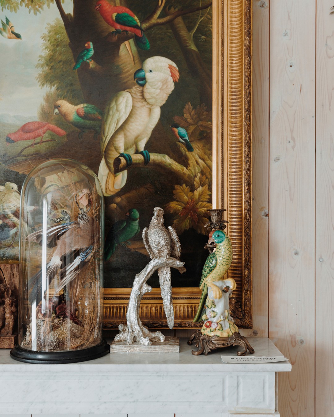 Artwork featuring a parrot painting, bird figurines, and a taxidermied bird under glass, all placed on a white mantelpiece.
