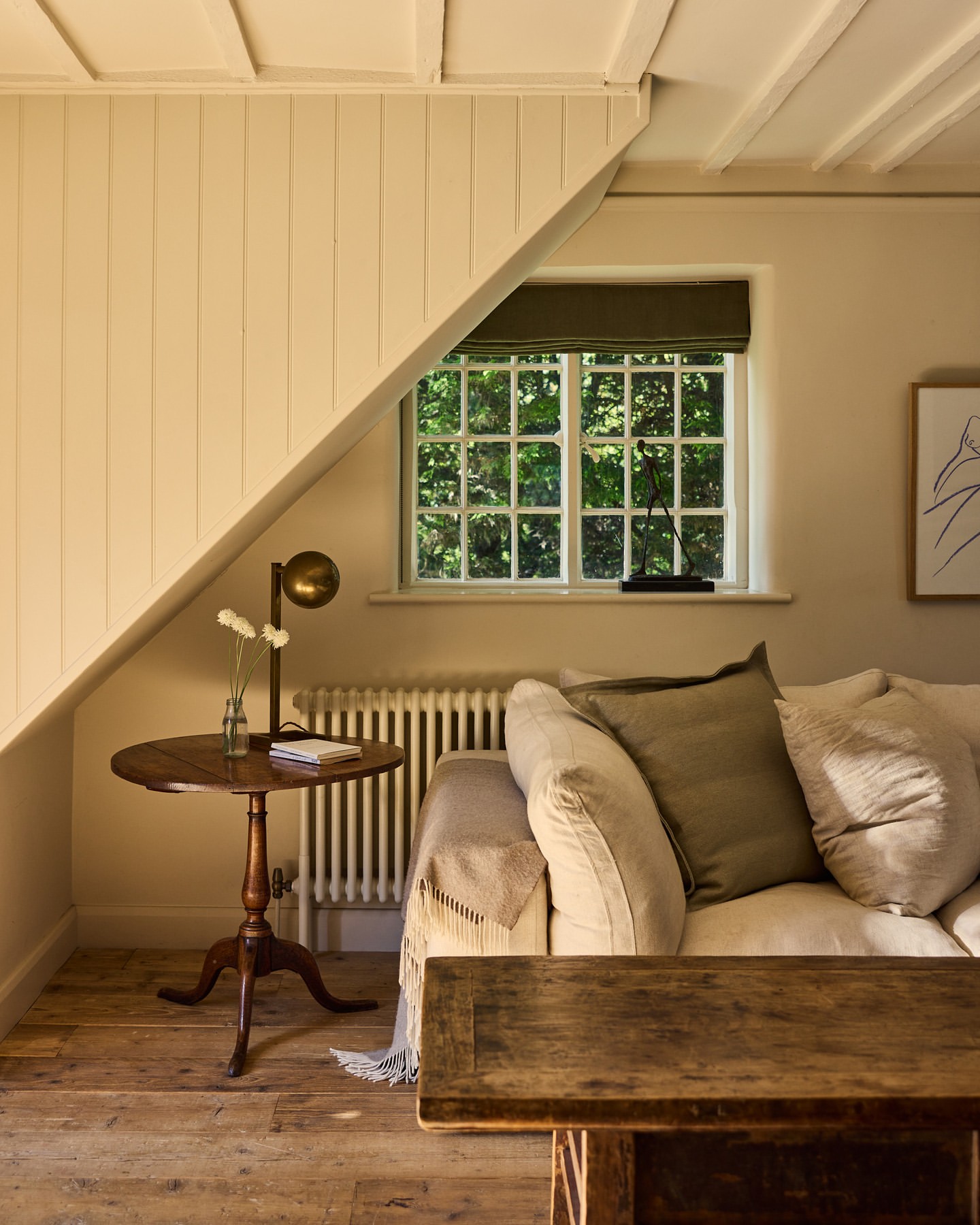 Cozy home interior features a cream sofa with pillows, a round wooden table with a lamp, and a window with natural light streaming in.