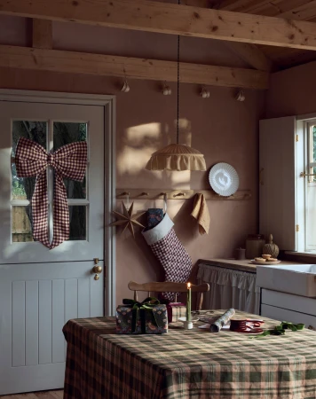 Holiday decor in a rustic kitchen: a plaid tablecloth and a large checkered bow on the door, accented by a stocking and gifts.