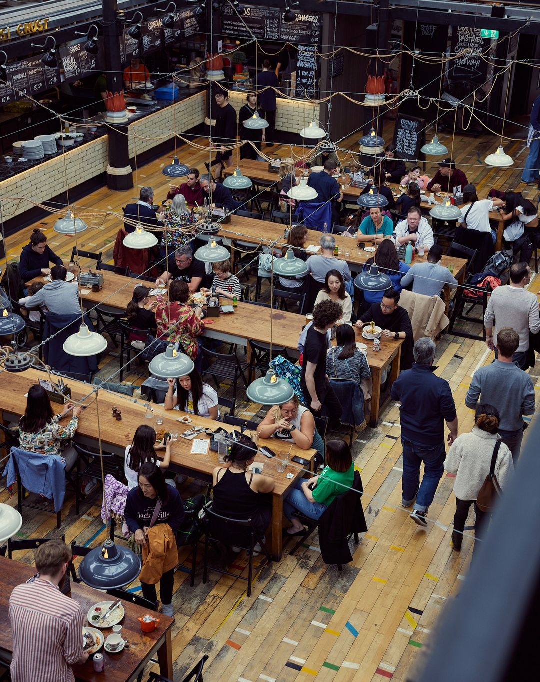 High-angle view of a bustling restaurant filled with people seated at wooden tables, illuminated by hanging pendant lights creating a warm ambiance.