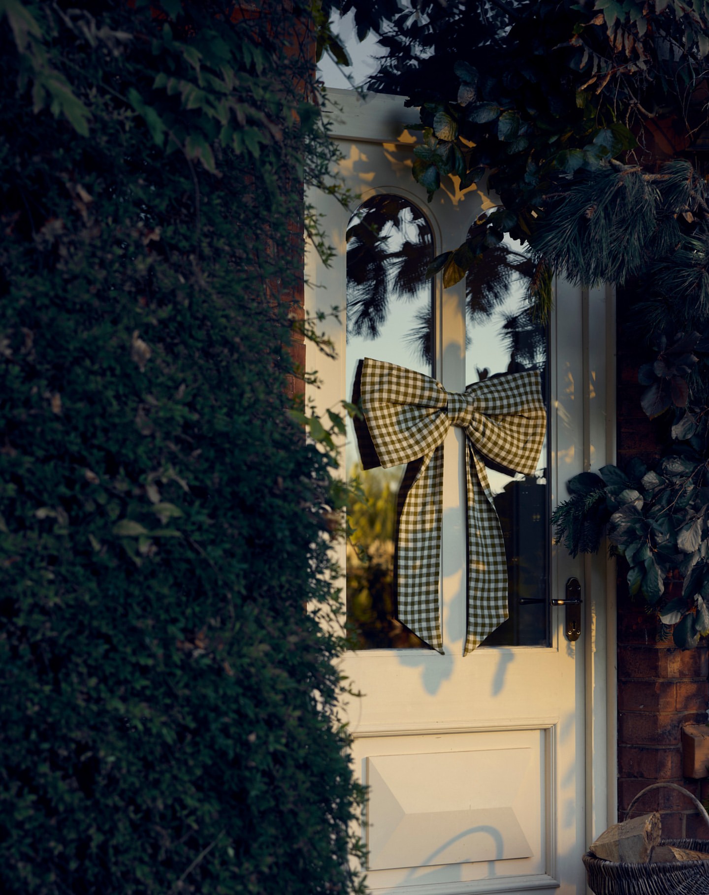 Front door adorned with a large, checkered bow, set against a backdrop of lush greenery and warm, inviting light.