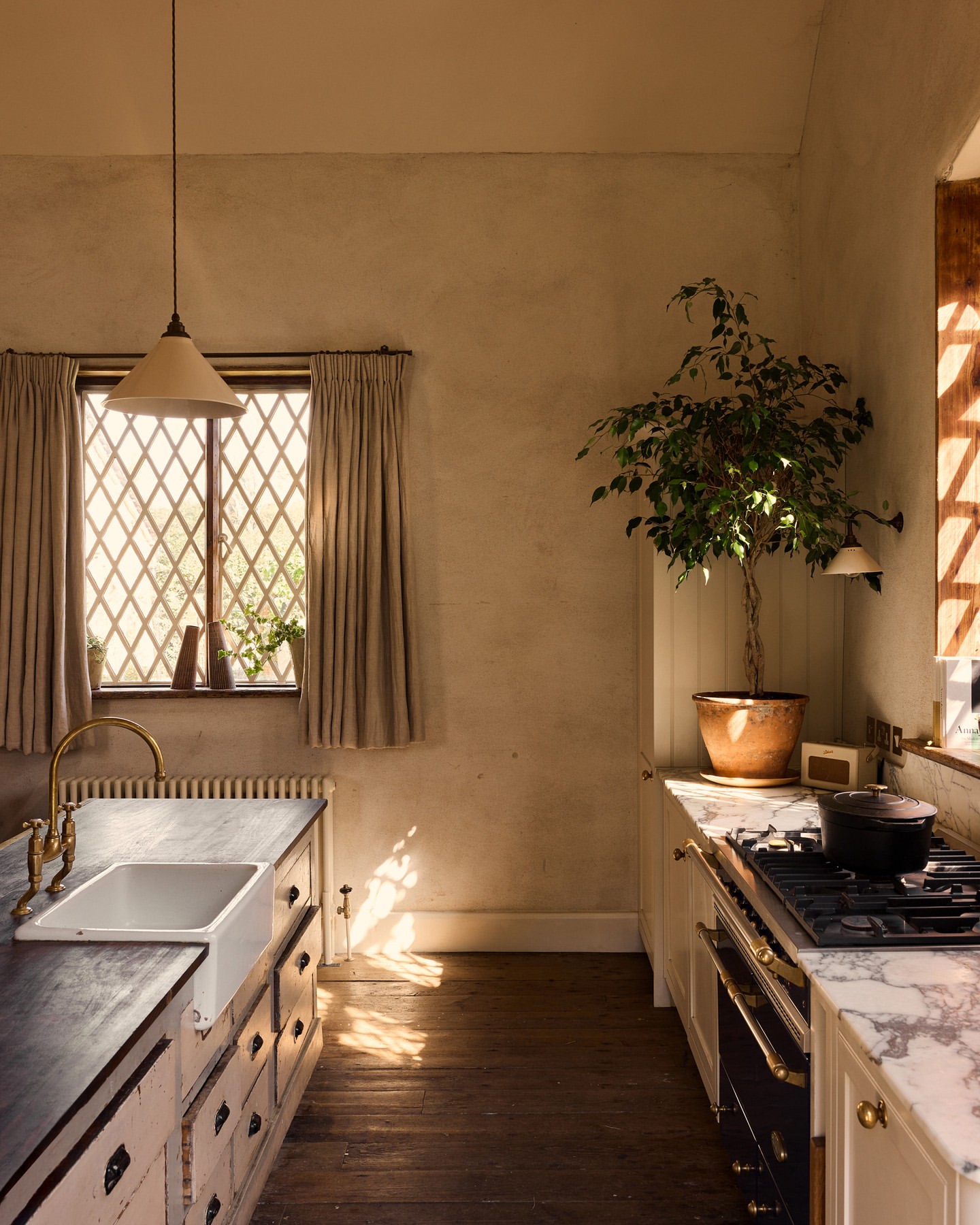 Sunlit kitchen interior with a white farmhouse sink, brass faucet, marble countertops, wooden floors, and a large potted plant.