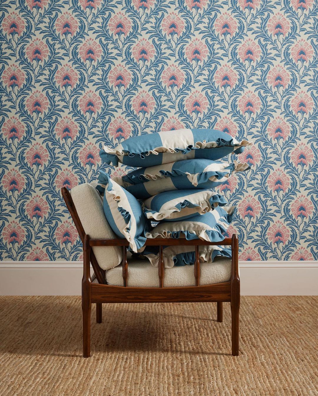 Interior scene with wooden chair stacked with striped blue and white ruffle pillows, floral patterned wallpaper, and jute rug.
