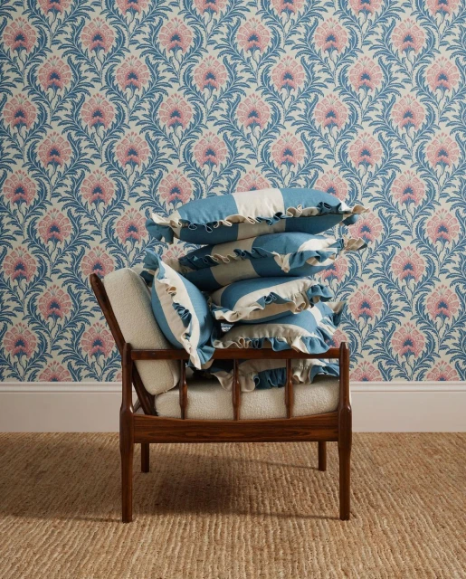 Interior scene with wooden chair stacked with striped blue and white ruffle pillows, floral patterned wallpaper, and jute rug.
