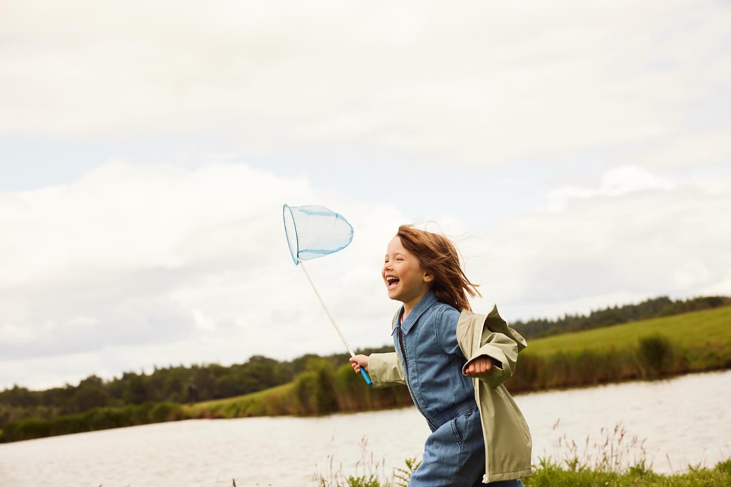 A child in a denim jumpsuit joyfully runs along the grassy bank of a lake, holding a blue net against a backdrop of sky and trees.
