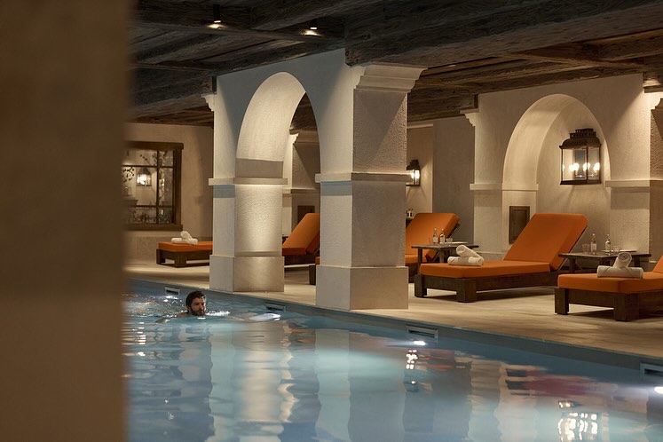 Indoor pool with a man swimming, orange lounge chairs with towels and tables with water bottles, architectural arches and wood ceiling.