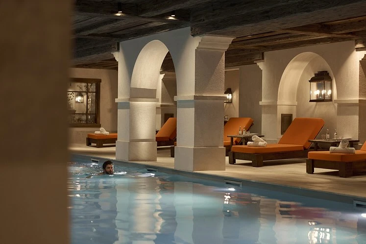 Indoor pool with a man swimming, orange lounge chairs with towels and tables with water bottles, architectural arches and wood ceiling.