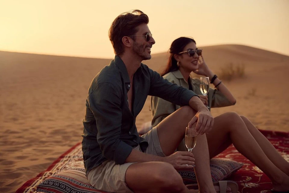 A stylish couple relaxes in the desert, seated on a patterned rug with pillows, enjoying champagne against a backdrop of sand dunes and sunset.
