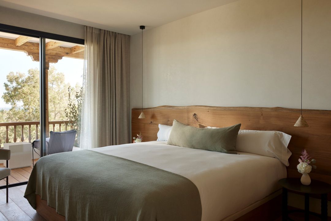 Bright bedroom interior showcasing a bed with a wooden headboard, bedside table, and a balcony with outdoor seating.
