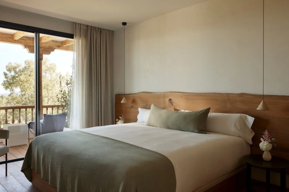 Bright bedroom interior showcasing a bed with a wooden headboard, bedside table, and a balcony with outdoor seating.