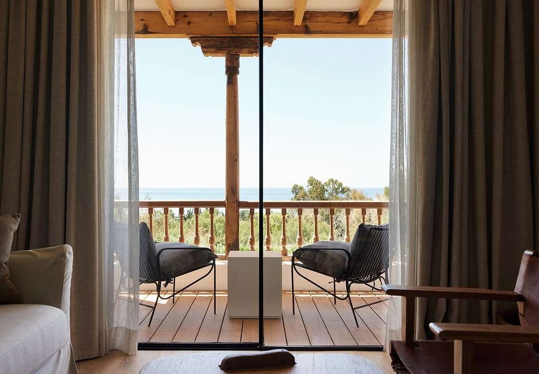 Interior view of a room with chairs on a balcony overlooking the ocean, framed by sheer curtains and a wooden structure.