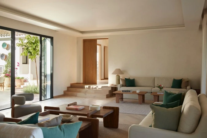 Interior view of a brightly lit living room with beige couches, wooden coffee tables, and view to an outdoor patio.