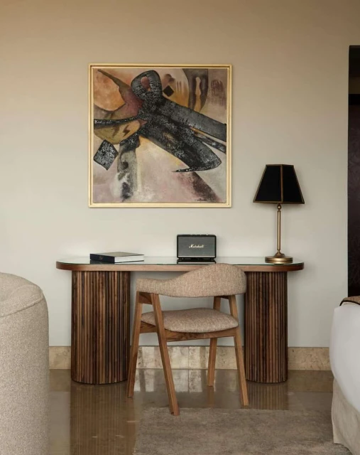 Interior shot featuring a desk with a painting above, a chair, a lamp, a speaker, and books against a neutral wall. Soft lighting.