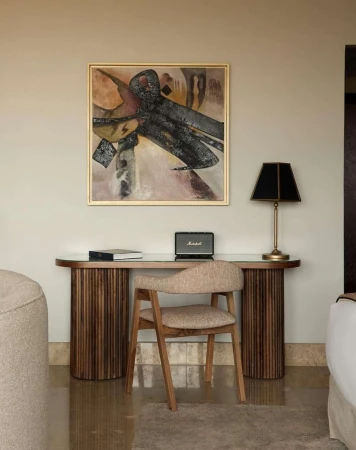 Interior shot featuring a desk with a painting above, a chair, a lamp, a speaker, and books against a neutral wall. Soft lighting.
