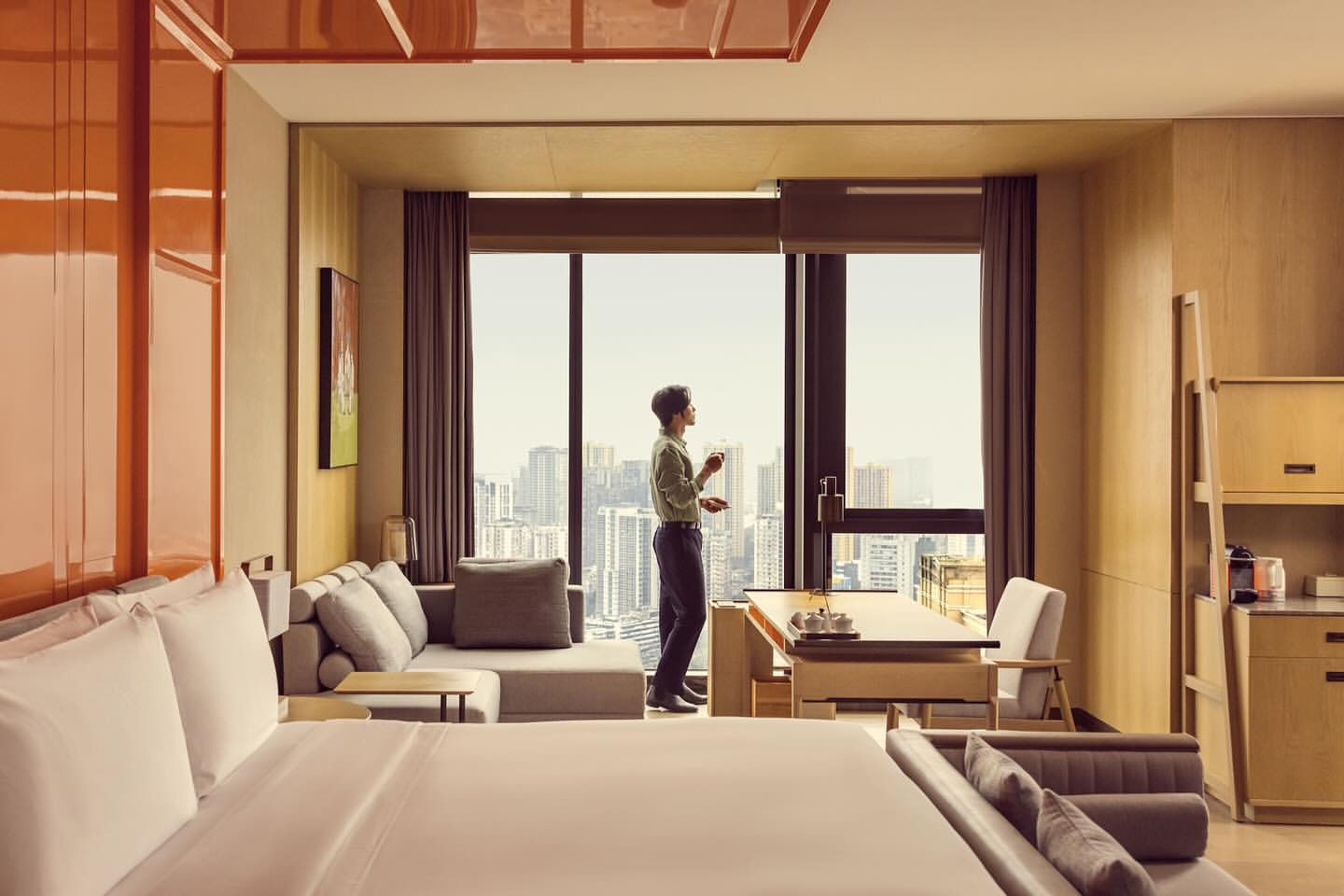 Hotel room interior featuring a man looking out the window, with a bed, seating area, and city view.