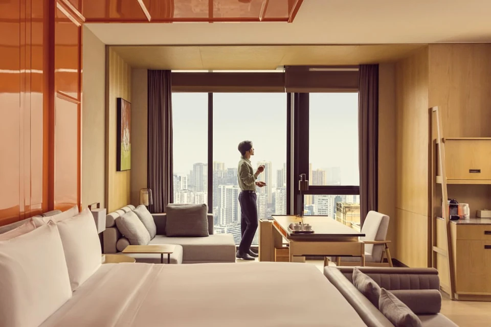 Hotel room interior featuring a man looking out the window, with a bed, seating area, and city view.