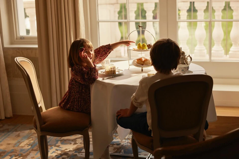 Young girl points at pastries on a tiered stand at a table, seated next to a child.  Indoor setting with natural window light.