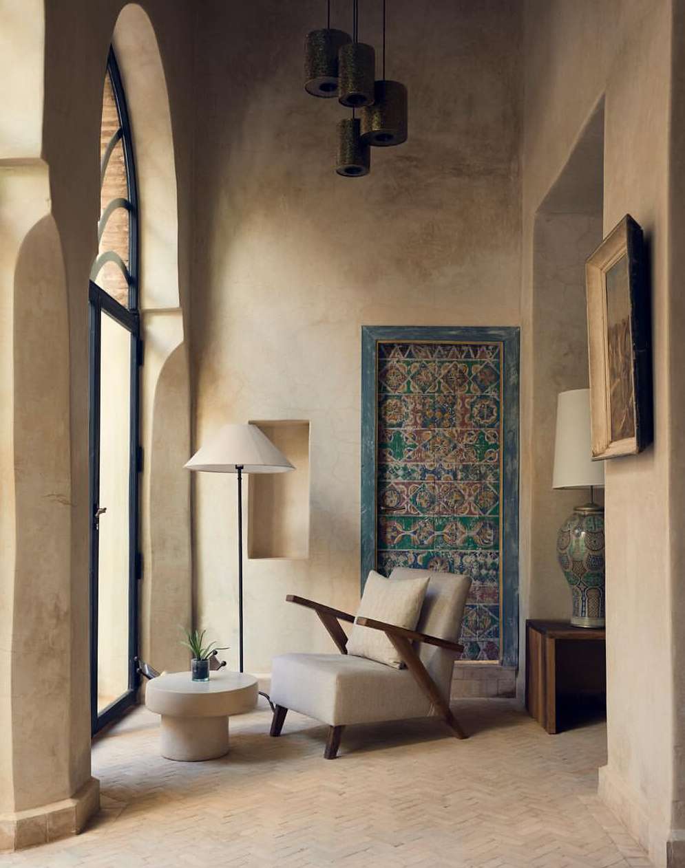 Indoor living space features a beige armchair, side table with plant, standing lamp, patterned doorway, and arched window.